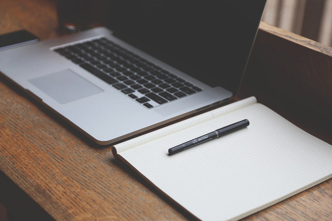 A laptop placed on a wooden table next to a notebook and pen.