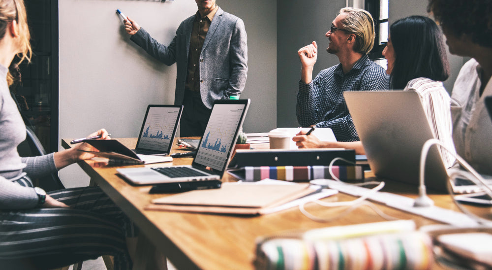 A team of people in a meeting with laptops on the table, while one person presents at a whiteboard.