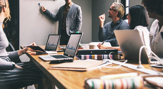 A team of people in a meeting with laptops on the table, while one person presents at a whiteboard.