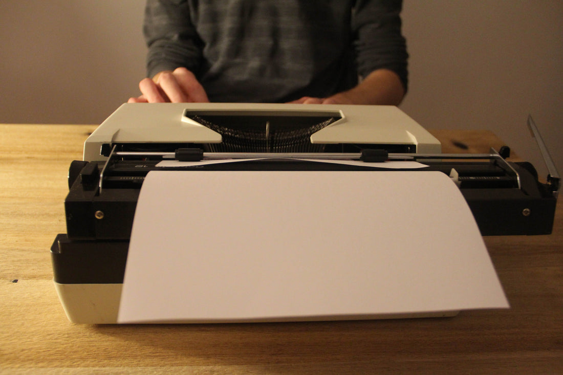 A typewriter with a blank sheet of paper, and hands visible in the background, suggesting typing.