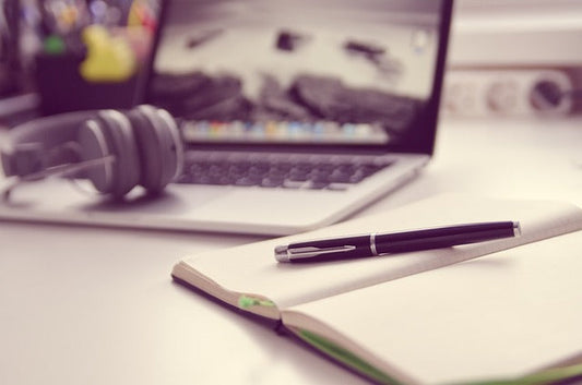 Pen and open notebook placed beside headphones and a laptop on a desk.