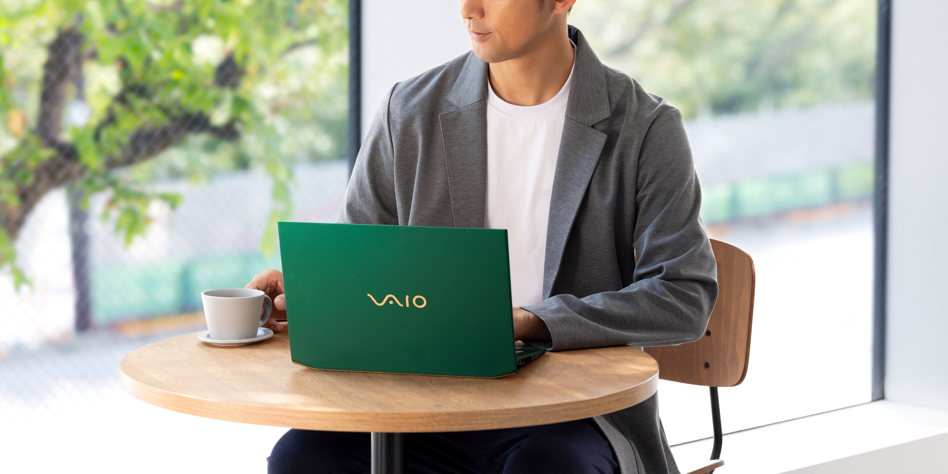 A man working on a green VAIO laptop at a café table, enjoying a cup of coffee with a bright outdoor view.