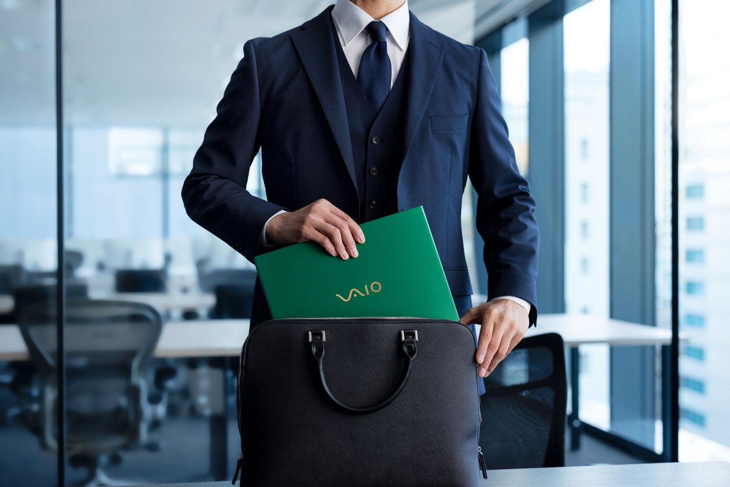 A businessman in a navy suit placing a sleek green VAIO laptop into a black leather bag in a modern office setting.