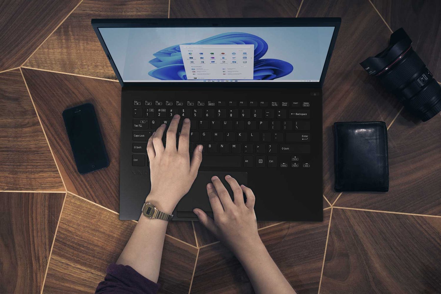 Person typing on a laptop with Windows 11, placed on a wooden table with a phone, wallet, and camera nearby.