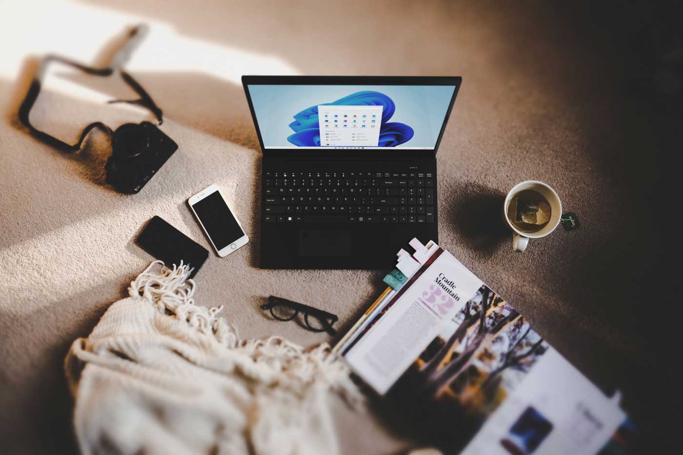 Laptop with Windows 11, camera, phones, glasses, magazine, coffee, and blanket arranged on a cozy floor setup.