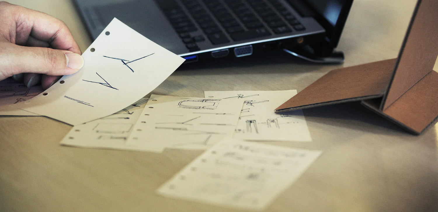 Hand holding sketch papers with design drawings next to a laptop and a folded cardboard model on a wooden desk.