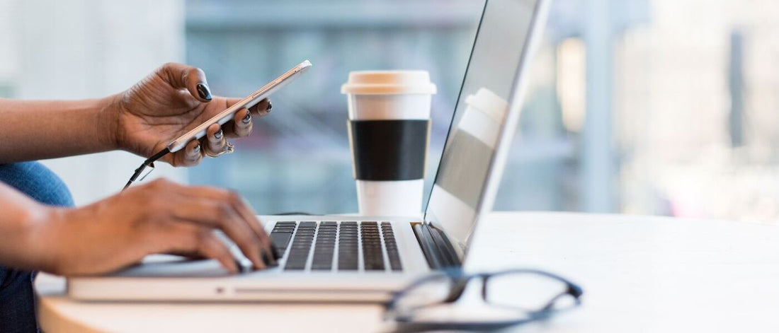 A person working on a laptop, holding a smartphone in one hand, with a coffee cup and glasses nearby.