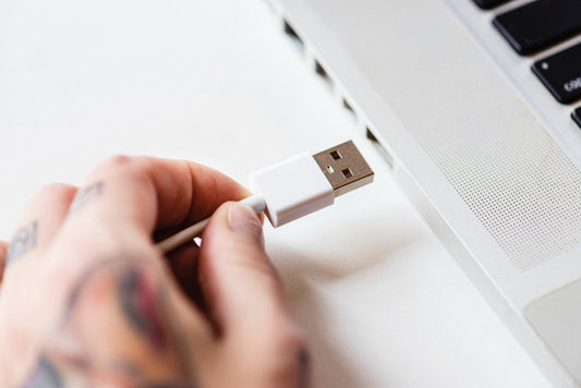 A person plugging a USB cable into a laptop. The hand has tattoos, and the laptop has a metallic finish.