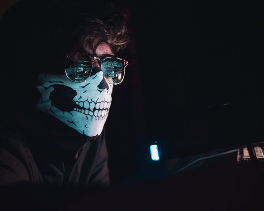 A person wearing a skull-patterned mask and sunglasses, working in low light, possibly at a computer.